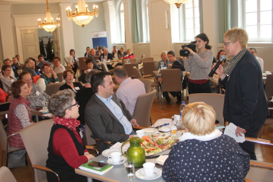 Menschen sitzen in einem Saal an Tischen, eine Person hält ein Mikrofon