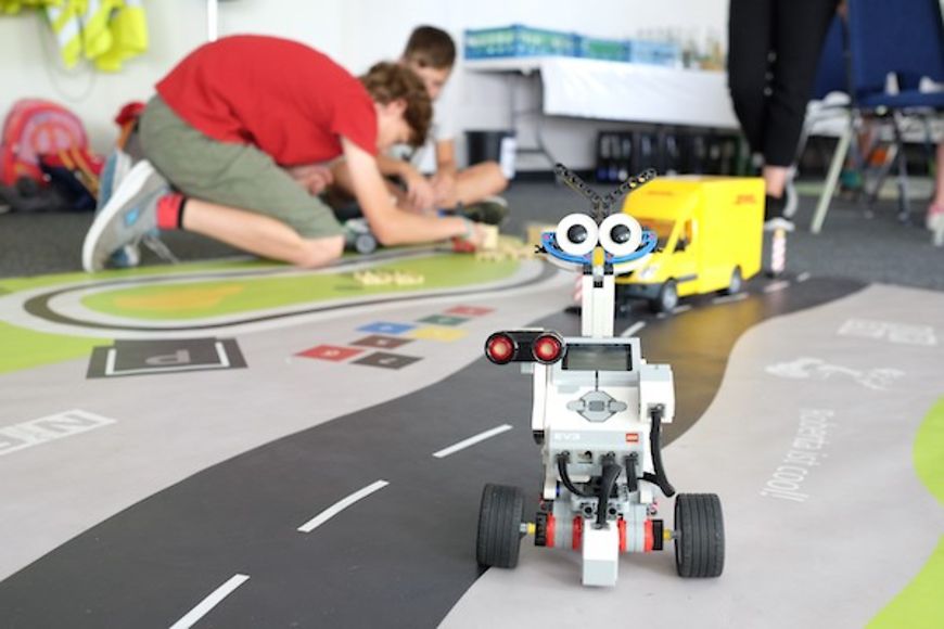 im Vordergrund ein Roboter aus Legotechnik, im Hintergrund spielende Kinder