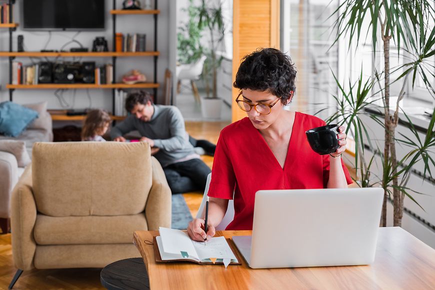 Eine Frau, die im Home Office arbeitet. Im Hintergrund beschäftigt der Vater das Kind. 