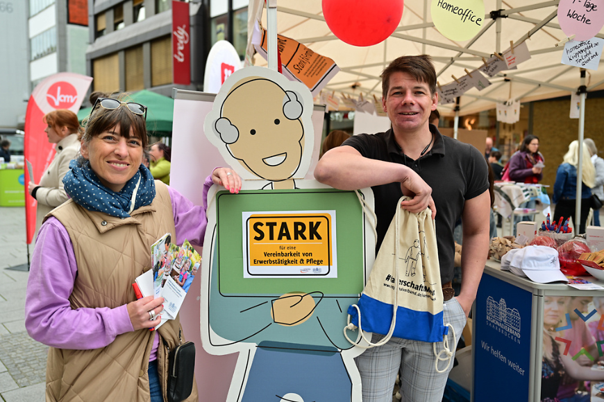 Man sieht zwischen einer Frau und einem Mann eine Figur aus Pappe, die für Vereinbarkeit von Beruf und Pflege wirbt. 